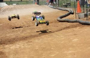 Three radio controlled cars come flying down an off-road track in a race. Two are airborne.